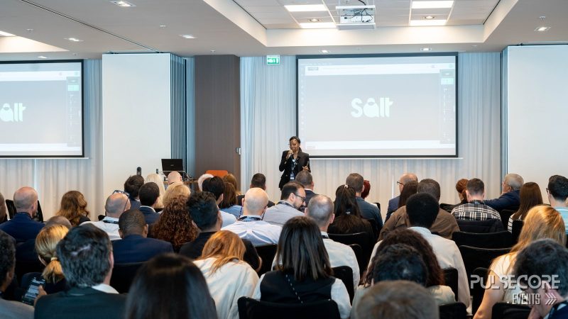 A full conference room facing the stage screens during a presentation at a previous PulseCore event