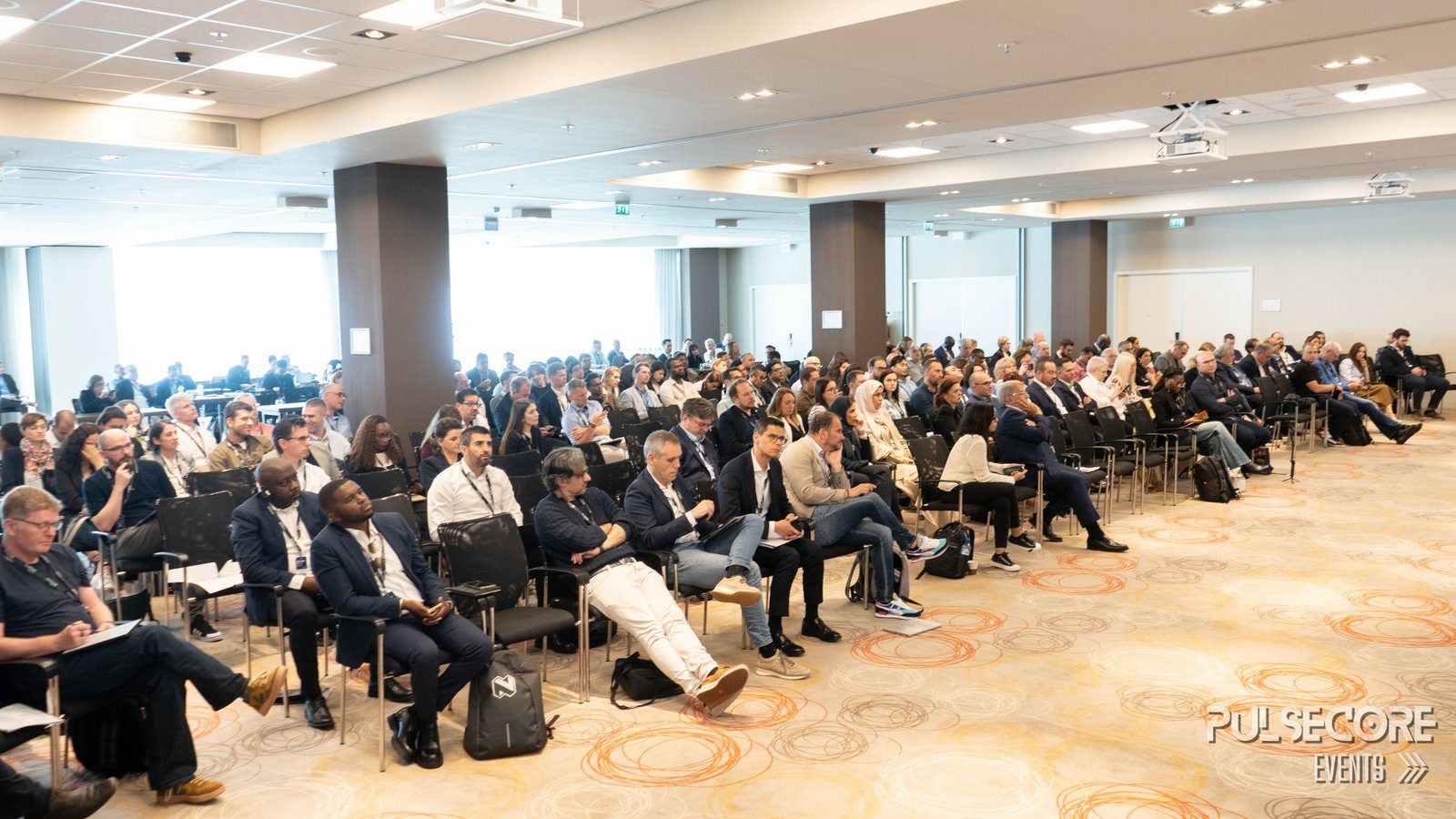 A wide view of a full conference hall, with rows of seated delegates and a daylight window wall behind them at a previous PulseCore event